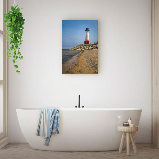 Photography wall art featuring the serene Crisp Point Lighthouse by a calm lake, with a clear blue sky above and smooth pebbles on the shore in the foreground.