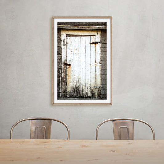 A captivating photography print featuring a beautifully weathered white door contrasting against the backdrop of aged grey shingles, inviting you into a world of rustic allure.